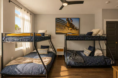 Sunlit modern bunk room in a vacation rental with two black metal bunk beds, blue geometric bedding and yellow throws, hardwood floors, ceiling fan, and horse-racing wall art.