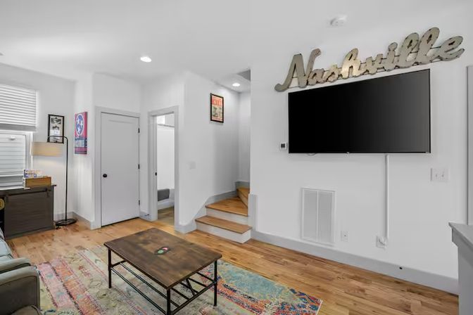 Bright, modern Nashville living room with wall-mounted TV beneath a metal “Nashville” sign, hardwood floors, colorful area rug, rustic coffee table, small desk by a window and a staircase leading upstairs.
