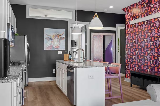 Contemporary open-plan kitchen with granite island and stainless-steel appliances, pendant lights, pastel pink bar stools, whimsical cow painting on a dark gray wall, purple cactus-print wallpaper and wood floors.