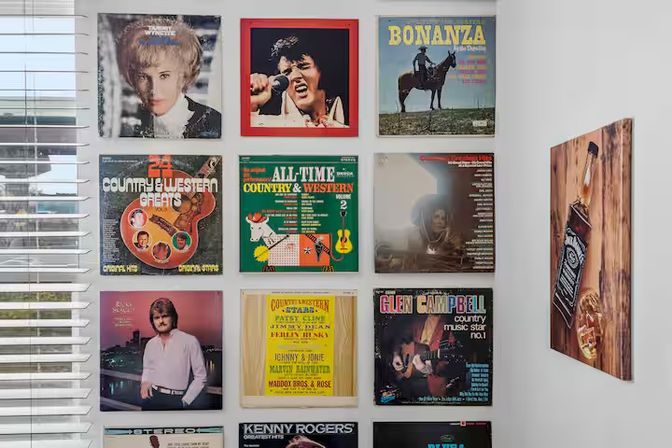 Retro country and western vinyl album covers in a neat grid on a white living-room wall beside a sunlit window, playful vintage music decor in a modern home