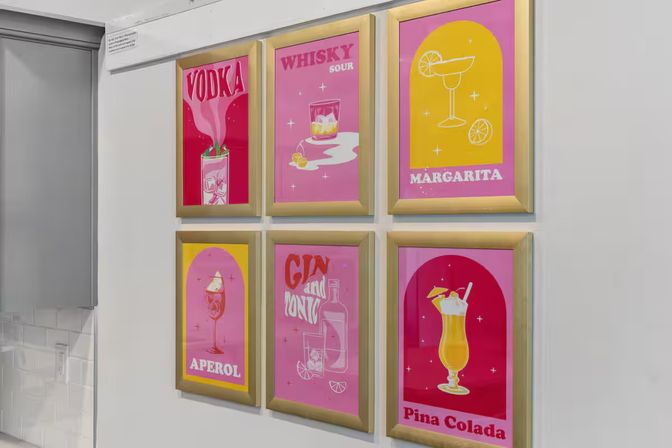 Playful 3x2 grid of framed cocktail prints (Vodka, Whisky Sour, Margarita, Aperol, Gin & Tonic, Pina Colada) on a white kitchen wall — bright home-bar decor.