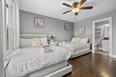 Bright cozy guest bedroom with two white-dressed beds, gray walls, hardwood floors, ceiling fan, window light and a small en-suite bathroom