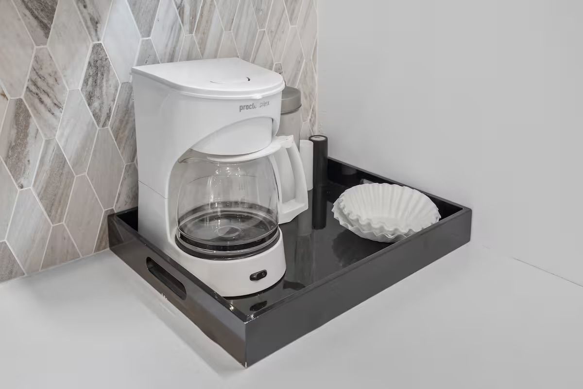 Compact kitchen coffee station with a white drip coffee maker and glass carafe on a black serving tray, paper filters beside it against a gray marble hexagon tile backsplash