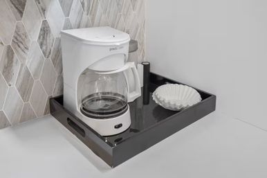 Compact kitchen coffee station with a white drip coffee maker and glass carafe on a black serving tray, paper filters beside it against a gray marble hexagon tile backsplash