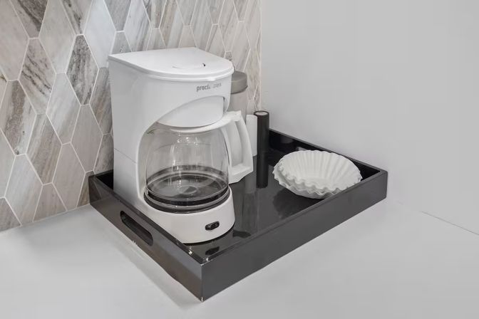 Compact kitchen coffee station with a white drip coffee maker and glass carafe on a black serving tray, paper filters beside it against a gray marble hexagon tile backsplash