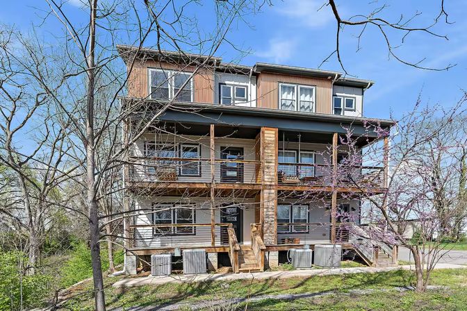 Three-story modern townhouse with wooden balconies and metal railings, front staircases and exterior HVAC units, framed by bare trees and a blooming purple tree on a sunny spring day in a suburban neighborhood