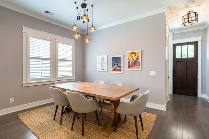 Modern dining room with a wooden farmhouse table and six upholstered mid-century chairs on a jute rug, gray walls with framed art, pendant cluster lights, hardwood floors and a dark wood front door.