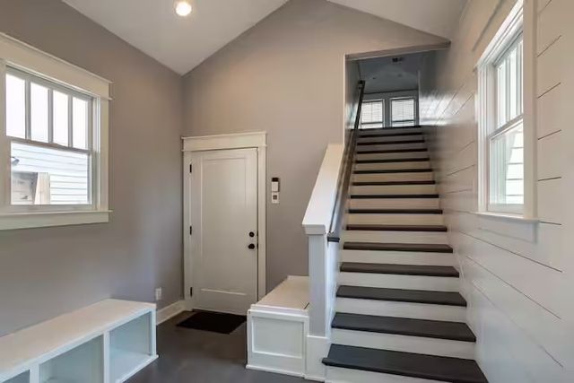 Bright modern home entryway with a straight staircase, white trim and shiplap wall, built-in bench with cubbies, neutral gray walls and windows letting in natural light.