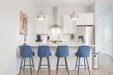 Sleek modern white kitchen with marble island, four blue velvet bar stools, stainless-steel fridge and range hood, pendant lights and colorful wall art.
