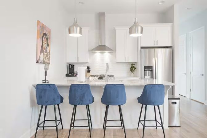 Sleek modern white kitchen with marble island, four blue velvet bar stools, stainless-steel fridge and range hood, pendant lights and colorful wall art.