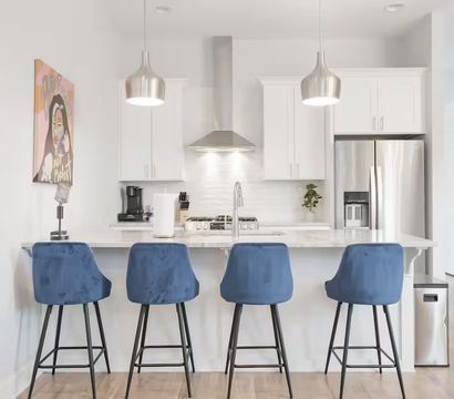 Sleek modern white kitchen with marble island, four blue velvet bar stools, stainless-steel fridge and range hood, pendant lights and colorful wall art.