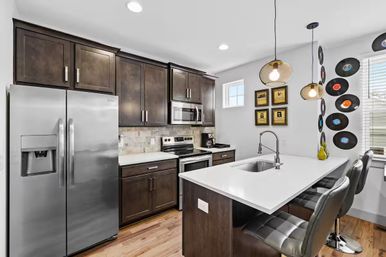 Bright modern kitchen with stainless-steel refrigerator and stove, dark wood cabinets, white quartz island with sink and three bar stools, pendant lights, hardwood floors and vinyl record wall decor.