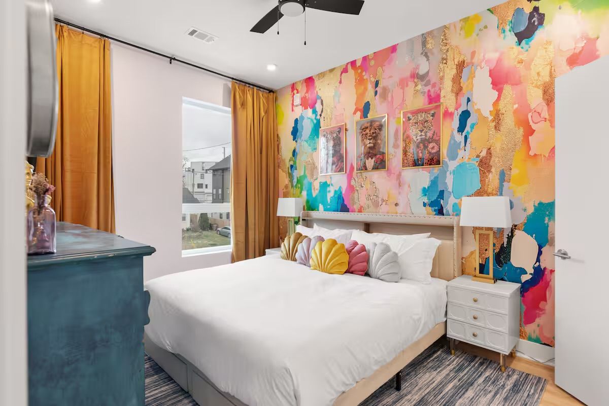 Vibrant city apartment bedroom with a colorful abstract watercolor-and-gold accent wall, framed animal prints, white bed with scallop-shaped pastel pillows, mustard curtains, white nightstands, teal dresser and ceiling fan.
