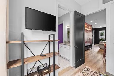 Modern apartment hallway with wall-mounted flat-screen TV above wood-and-metal shelves (remote and wire baskets), open black door to a bathroom with white vanity and purple accent wall, and view into a living room with patterned wall and hardwood floors.