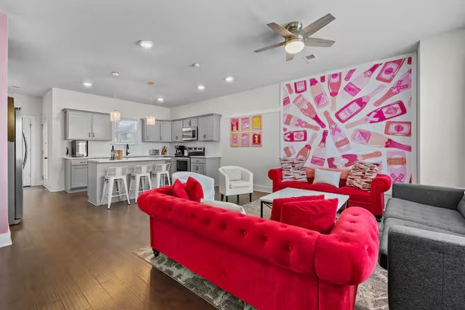 Open-plan modern living room and kitchen with bold pink champagne-bottle mural, red velvet tufted sofas, gray cabinets, white island with stools, and dark hardwood floors