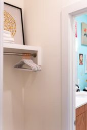 Small modern linen closet with white plastic hangers on a brass rod, folded white towels and gold-accent framed art, open to a turquoise-walled bathroom with wooden vanity.