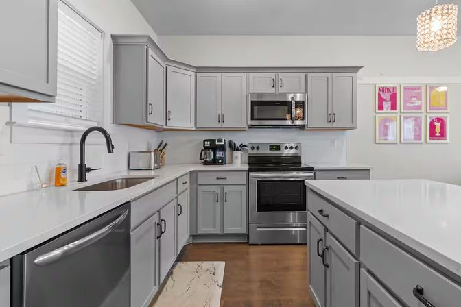 Bright modern gray kitchen with shaker cabinets, white quartz countertops, stainless steel stove, microwave and dishwasher, large island, black gooseneck faucet, hardwood floors and colorful framed cocktail prints on the wall.