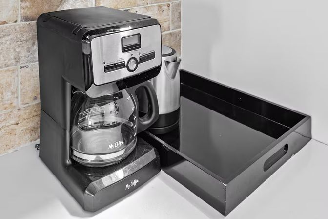 Sleek black drip coffee maker with glass carafe on a white kitchen countertop, forming a compact coffee station next to a black serving tray and stainless milk frother against a tiled backsplash.