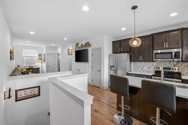 Open-plan Nashville apartment interior with sleek dark-wood kitchen cabinets, stainless-steel appliances, marble tile backsplash, breakfast bar with two black stools, hardwood floors and a cozy living area