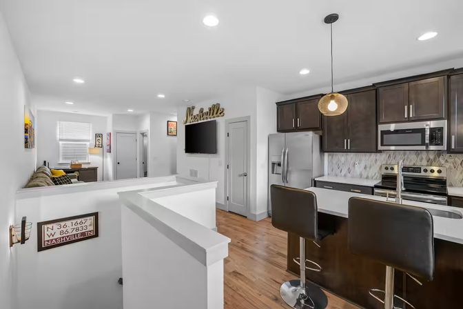 Open-plan Nashville apartment interior with sleek dark-wood kitchen cabinets, stainless-steel appliances, marble tile backsplash, breakfast bar with two black stools, hardwood floors and a cozy living area