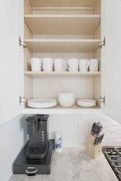 Open white kitchen cabinet with neatly arranged white mugs and stacked plates above a marble countertop coffee station featuring a single‑serve brewer, creamer canister and wooden knife block beside subway tile backsplash.