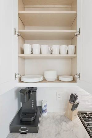Open white kitchen cabinet with neatly arranged white mugs and stacked plates above a marble countertop coffee station featuring a single‑serve brewer, creamer canister and wooden knife block beside subway tile backsplash.