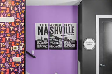 Three-panel NASHVILLE city skyline wall art on a bright purple entry wall, flanked by playful western wallpaper with cacti, cowboy boots and skulls on the left and a dark gray door with a round sign on the right; thermostat, light switches and a stair handrail visible.