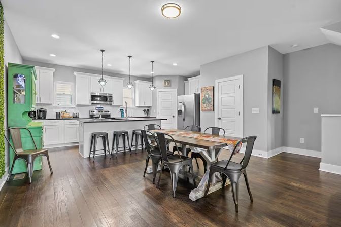 Bright open-plan kitchen and dining room with white shaker cabinets, island with metal bar stools, rustic farmhouse wood table surrounded by industrial metal chairs, green accent hutch, stainless refrigerator and dark hardwood floors.