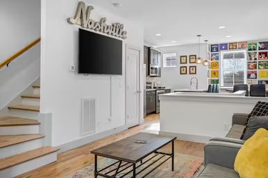Open-concept Nashville living room and kitchen with wall-mounted TV beneath a 'Nashville' sign, wooden staircase, gray sofa with yellow pillow, rustic coffee table, and colorful gallery wall.