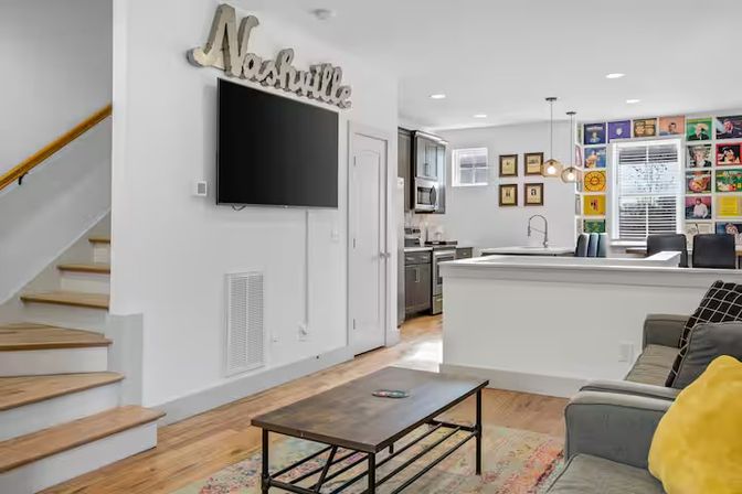 Open-concept Nashville living room and kitchen with wall-mounted TV beneath a 'Nashville' sign, wooden staircase, gray sofa with yellow pillow, rustic coffee table, and colorful gallery wall.