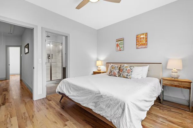 Bright contemporary bedroom with white bedding, floral accent pillows, wooden nightstands and lamps, ceiling fan, en-suite glass shower and warm hardwood floors