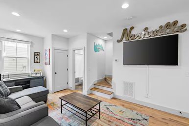 Bright modern Nashville-themed living room with gray sectional sofa, wooden coffee table on a colorful area rug, wall-mounted flat-screen TV beneath a 'Nashville' sign, hardwood floors and a short staircase.