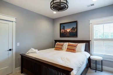 Cozy guest bedroom with dark wooden bed frame, crisp white linens and folded towels, colorful patterned pillows, gray walls, framed city sunset print and shuttered window.