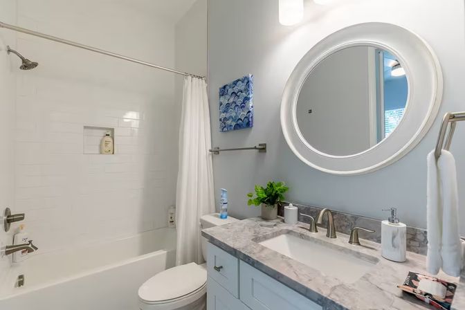 Bright, spa-like modern bathroom with marble countertop vanity, round mirror, brushed-nickel faucets, white subway-tile bathtub with shower curtain, toilet and small potted plant.
