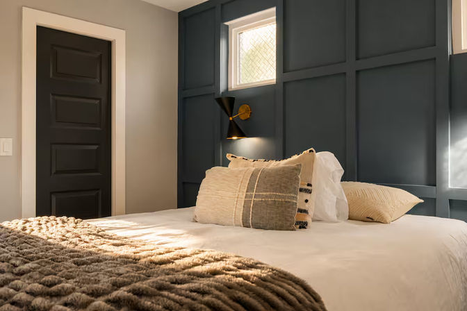 Cozy modern bedroom interior with navy paneled accent wall and small frosted window, brass wall sconce over a neatly made bed with textured beige pillows and a chunky knit throw blanket bathed in sunlight.