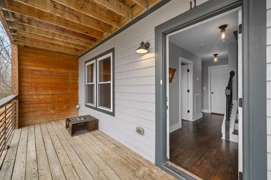 Cozy covered front porch with weathered wooden plank floor, wood-paneled wall and ceiling, metal railing and exterior wall light opening into a home with dark hardwood floors, modern light fixtures and a staircase.