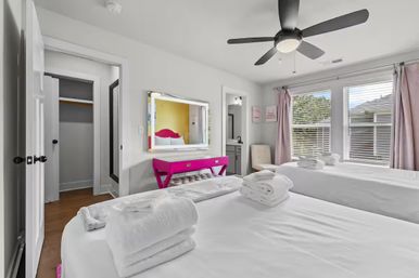 Bright residential twin bedroom with two beds in crisp white linens and folded towels, sunlit windows with blush curtains, black ceiling fan, hot-pink vanity with mirror, neutral walls and hardwood floor.