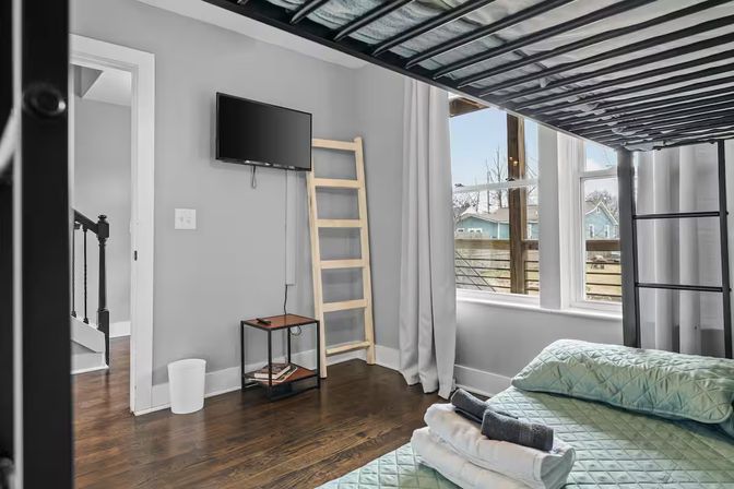 Sunlit bedroom with metal bunk bed and mint-green quilted lower bed topped with folded towels, wall-mounted TV, wooden ladder decor, small side table and hardwood floors beside a large window overlooking neighboring houses.