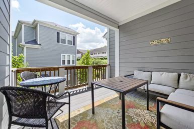 Covered suburban townhouse balcony patio with gray siding, wicker bistro table and chairs, cushioned L-shaped outdoor sofa, wooden coffee table on a floral rug, wooden railing, neighboring gray houses visible and a small “Please No Smoking” sign on the wall.