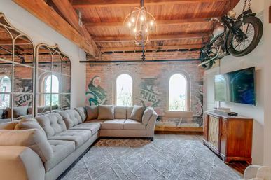 Sunlit industrial-chic loft living room with exposed brick and arched windows, wood-beam ceiling, tufted gray sectional sofa, mirrored accent wall, patterned area rug, wall‑mounted vintage motorcycle and flat-screen TV.