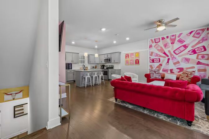 Modern open-plan living area and kitchen with a bold pink bottle-print accent wall, red velvet tufted sofa, white island with three bar stools, gray cabinets, hardwood floors and ceiling fan — contemporary apartment interior.