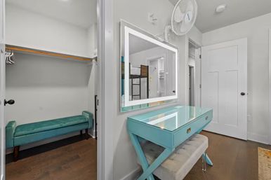 Bright bedroom dressing nook with a turquoise glass-top vanity and tufted bench beneath an illuminated LED mirror, adjacent walk-in closet with teal bench and wooden hangers, white walls and hardwood floors.