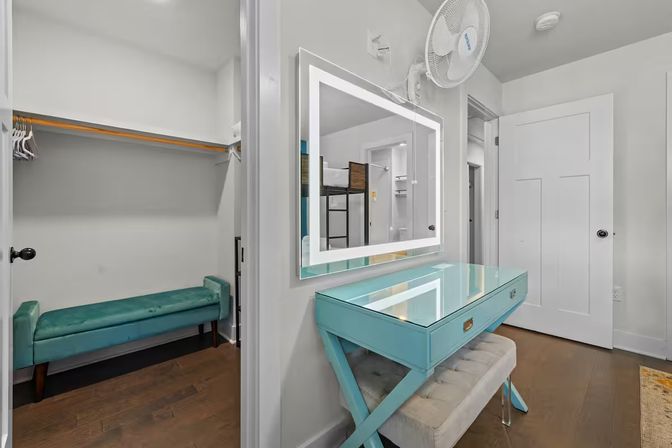 Bright bedroom dressing nook with a turquoise glass-top vanity and tufted bench beneath an illuminated LED mirror, adjacent walk-in closet with teal bench and wooden hangers, white walls and hardwood floors.