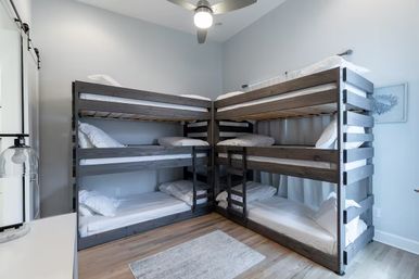 L-shaped bedroom with two three-tier dark wood bunk beds (six single beds), white linens, light gray walls, hardwood floor, ceiling fan and small area rug, sleepover-ready.