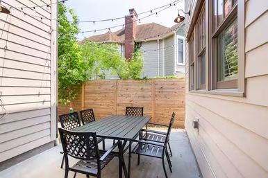 Cozy narrow backyard patio with a metal outdoor dining table and six chairs, overhead string lights, wooden privacy fence, concrete floor, and neighboring houses visible
