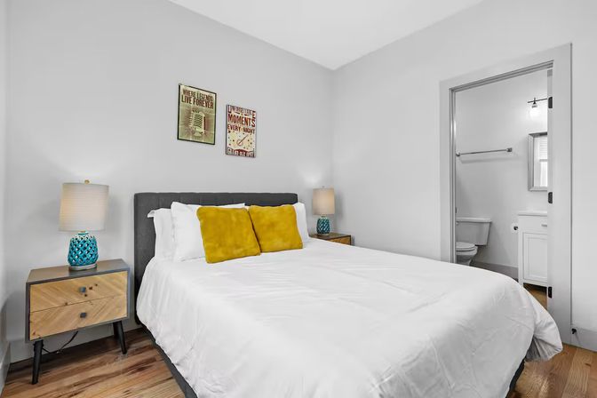 Bright modern bedroom interior with white bedding and gray headboard, mustard yellow accent pillows, wooden nightstands with turquoise lamps, hardwood floors and doorway to en-suite bathroom.