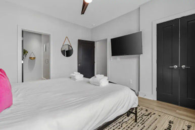 Stylish modern apartment bedroom with large white bed, folded towels, pink accent pillow, wall-mounted TV, round hanging mirror and visible en-suite glass shower, light wood floor and black doors.