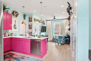 Vibrant open-concept kitchen and living room with hot-pink lower cabinets, aqua walls, white island with stainless dishwasher, gold pendant lights, floral accent chair, colorful artwork and a large window overlooking trees.