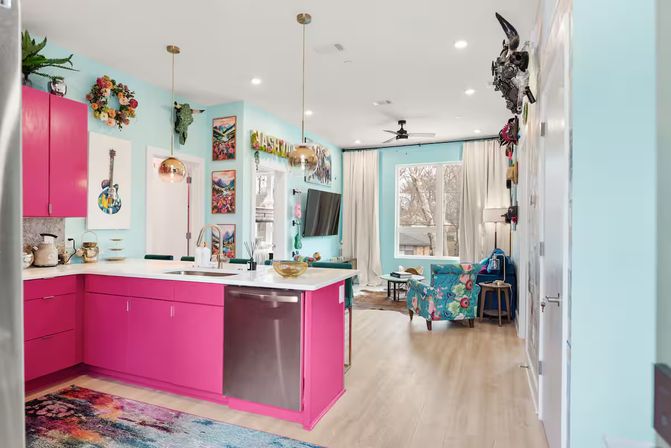 Vibrant open-concept kitchen and living room with hot-pink lower cabinets, aqua walls, white island with stainless dishwasher, gold pendant lights, floral accent chair, colorful artwork and a large window overlooking trees.