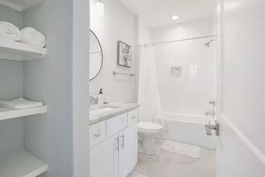 Bright modern white bathroom interior with marble-topped vanity and round mirror, open shelves with folded towels, toilet and tub-shower with white curtain